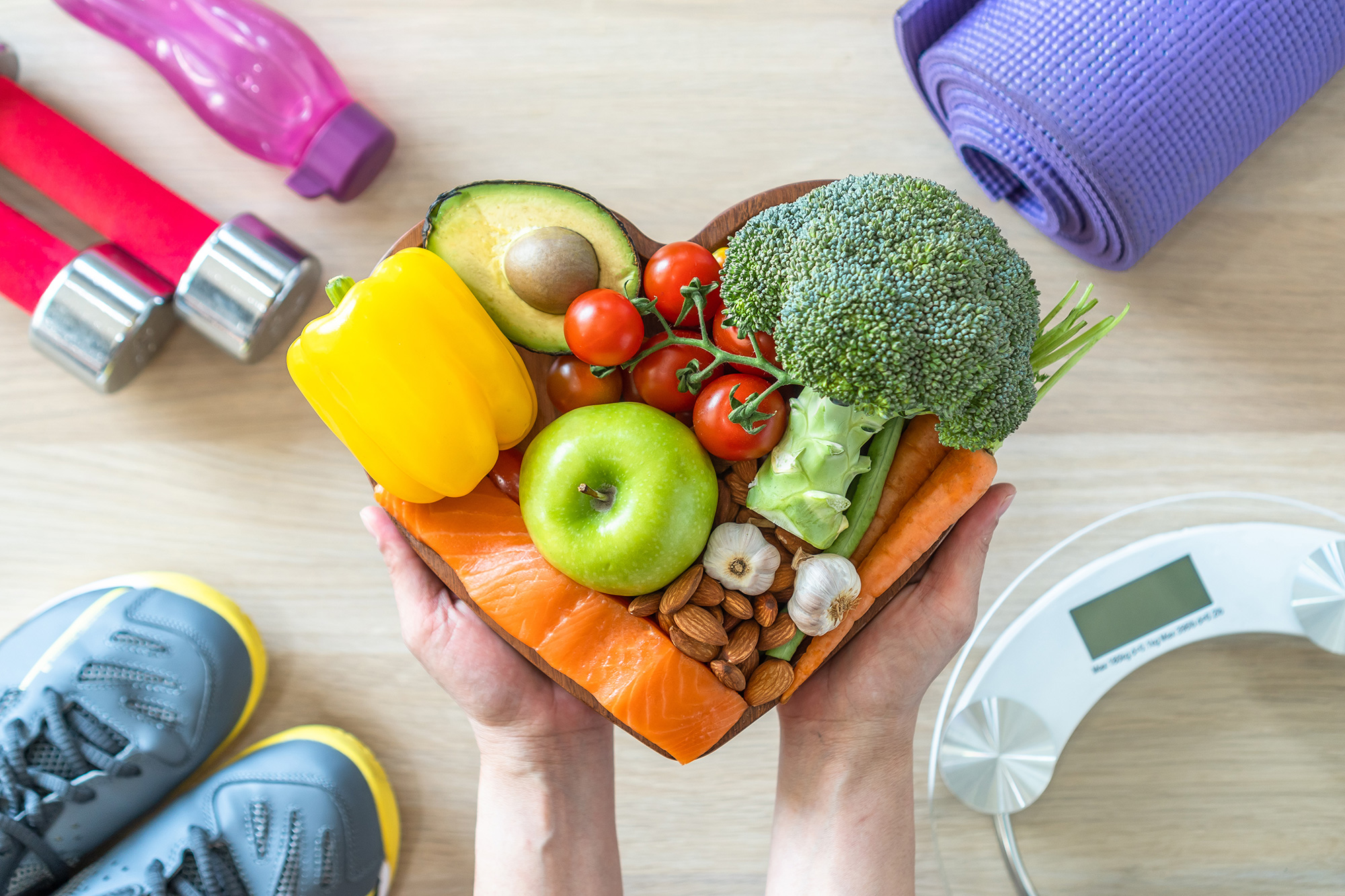 Photo of person holding heart-shaped bowl of whole foods. The bowl is surrounded by sports shoes, exercise equipment, and scales.